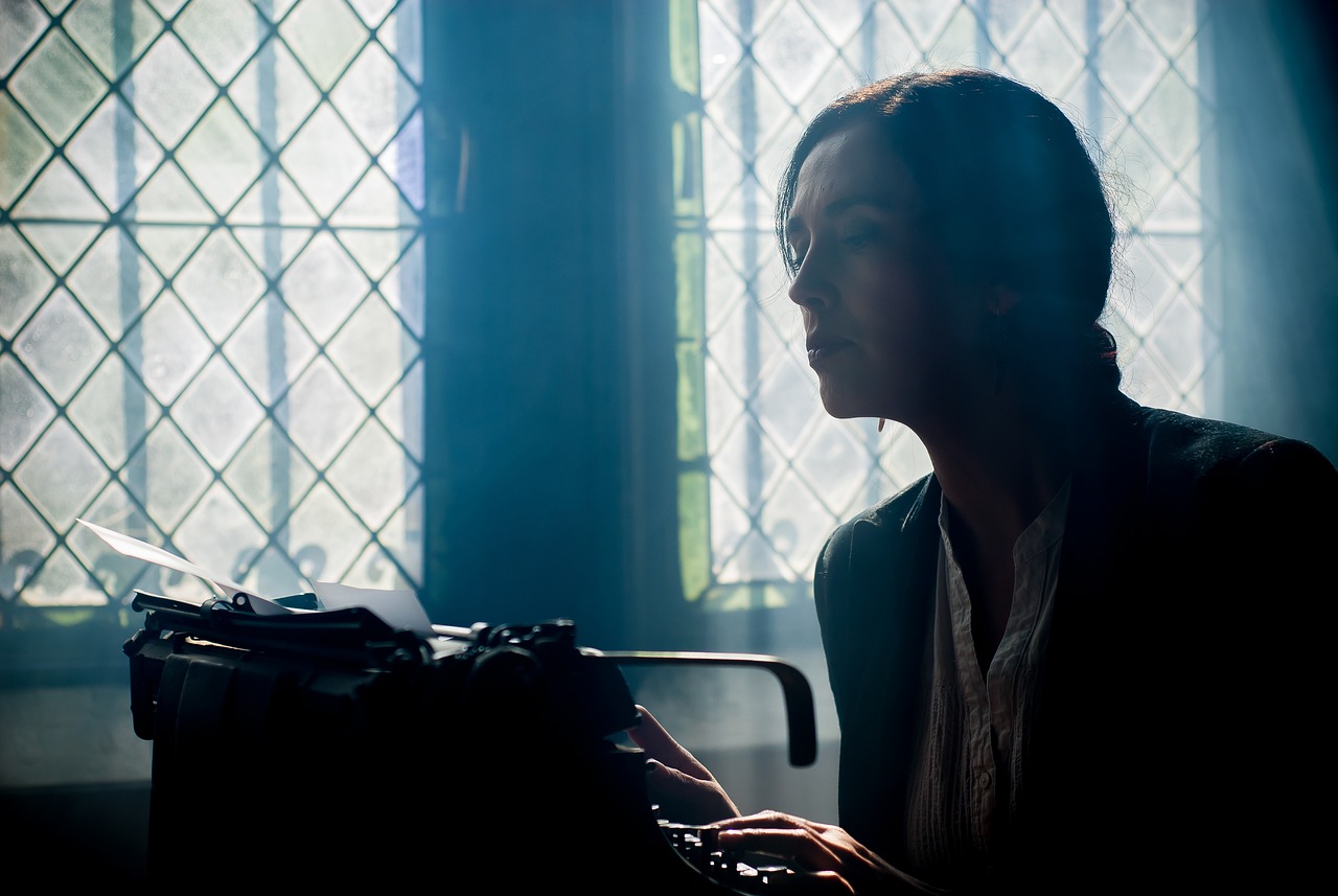 Stock image of someone tapping at a typewriter in a dark room by the window.