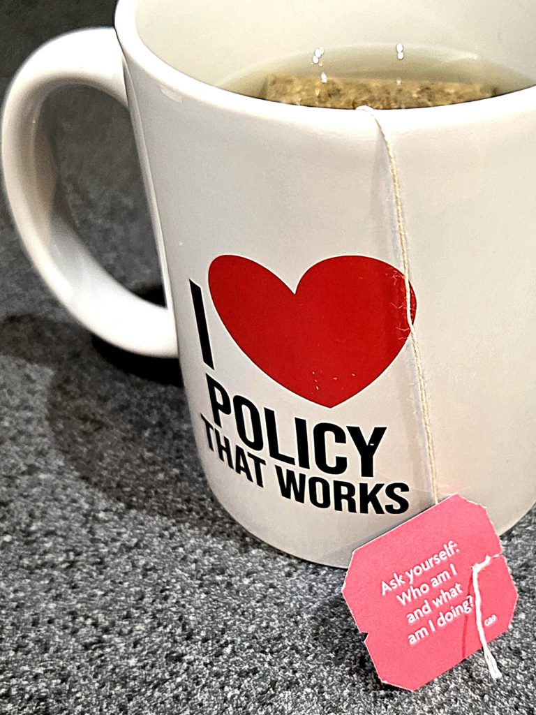 A white tea-filled mug reads “I ❤️ POLICY THAT WORKS,” with a tea tag asking, “Who am I and what am I doing?”